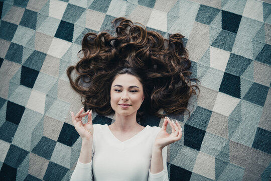 Young woman lying on patterned rug with long hair, relaxed and meditating in a calm cozy home top-view portrait