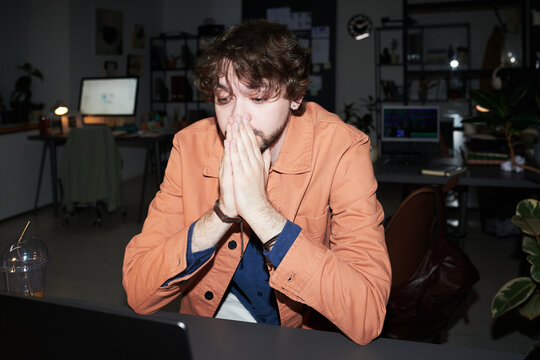Young adult Caucasian man covering mouth while staring at laptop in office, showing stress after work issue. Useful for burnout, deadline pressure, remote work crisis marketing