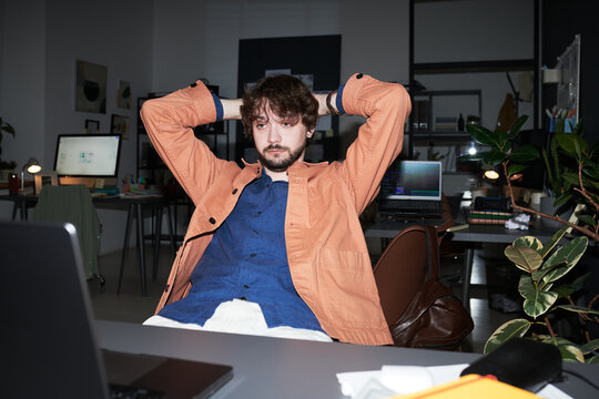 Young adult Caucasian man leaning back with hands behind head, looking at laptop in office, reflecting on workload. Useful for remote work, burnout, productivity themes