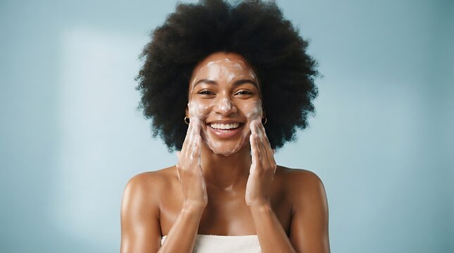 Cheerful black female model applies foaming cleanser, has clean fresh healthy skin, smiles broadly, Afro bushy haircut, stands bare shoulder against blue background. Beauty and feminine concept