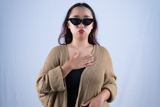 Young Asian woman in brown cardigan and sunglasses with hand on chest on white background