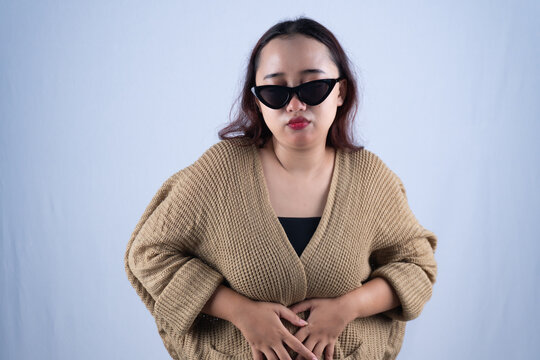 Young Asian woman in sunglasses holding stomach with pain against white background