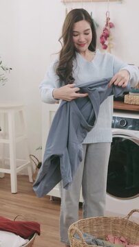 A smiling Asian woman doing laundry at home. She smells the fresh, clean clothes and hangs a t-shirt on a wooden clothes rack.