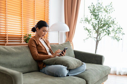 Happy young asian woman relax on comfortable couch at home texting messaging on smartphone, smiling girl use cellphone, chatting online message, shopping online from home