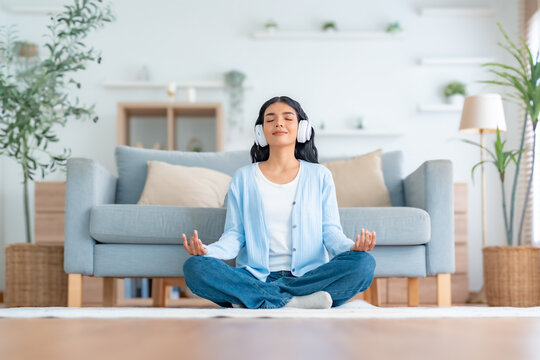 Happy young Asian woman practicing yoga and meditation at home sitting on floor in living room in lotus position and relaxing with closed eyes. Mindful meditation and wellbeing concept