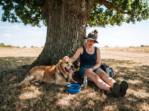 Happy woman resting under a large tree with her golden retriever dog on a sunny summer day