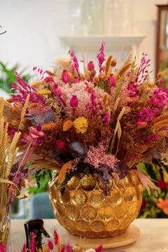 Elegant arrangement of dried flowers, preserved botanicals and ornamental grasses displayed in a decorative golden vase inside a flower shop. 