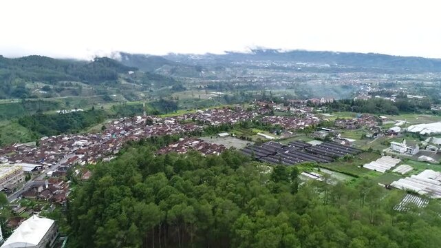 Aerial drone footage flying forward over a residential settlement near Tangkuban Parahu, West Java, Indonesia, showing dense rooftops, greenery, and rolling hills with misty mountains in the backgroun