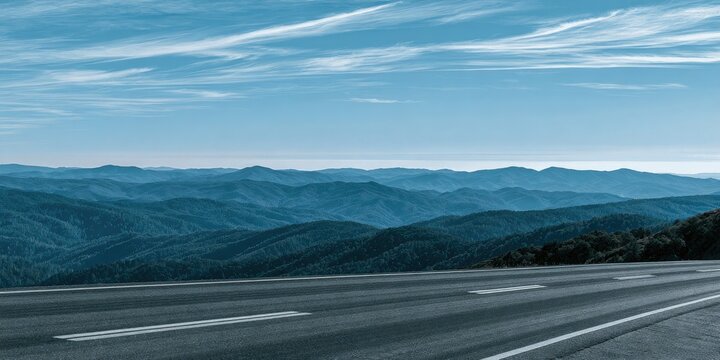 An empty asphalt road curves through a vast expanse of blue, mist-shrouded mountains under a cloudy sky
