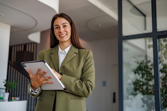 Businesswoman standing in modern office holding a tablet, smiling and looking at camera while working