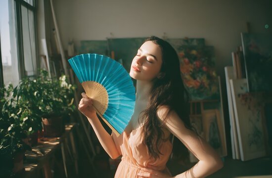 Plan Moyen d une Jeune Femme se Rafra&icirc;chissant avec un &Eacute;ventail Bleu dans un Studio Ensoleill&eacute;