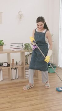 Playful young Asian woman in an apron and rubber gloves dancing and having fun with a mop while cleaning the floor.