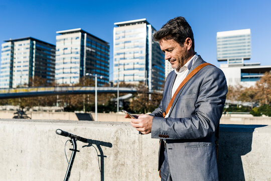 Mature businessman commuting in the city with his kick scooter, usine smartphone