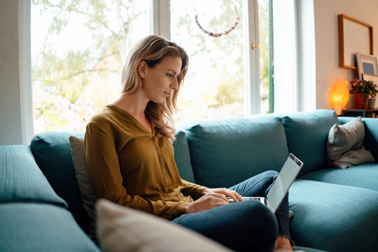 Blond woman using laptop sitting on sofa in living room at home