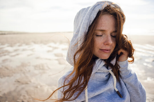 Netherlands, Zeeland, portrait of redheaded woman with freckles wearing hooded jacket on the beach