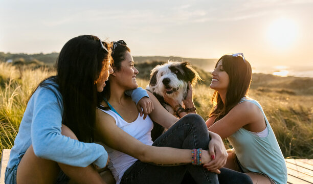 Three happy women with dog sitting on boardwalk in dunes at sunset