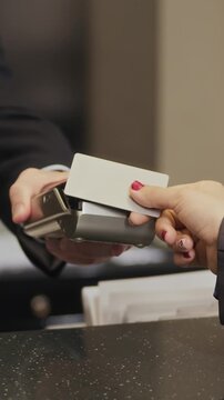 Vertical closeup of customer tapping blank credit card on payment terminal held by receptionist at hotel desk. Footage illustrates contactless payment methods and modern banking technology