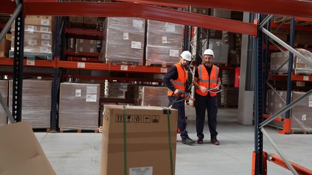 Two male warehouse employees in safety gear using a hand pallet truck to move a large cardboard box through a logistics distribution center