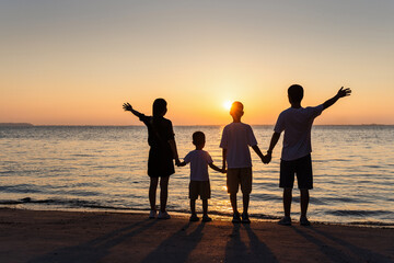 Fototapeta premium A family group of five stands on a beach at sunset, holding hands, with arms raised against the glowing sky and calm sea.