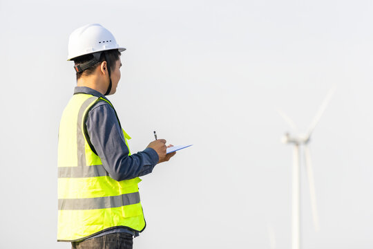 A male engineer wearing safety gear takes notes while inspecting a wind turbine at an outdoor power site.