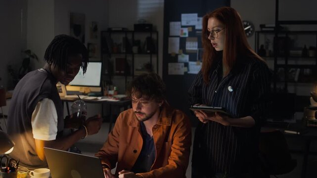Medium shot of diverse young adult colleagues, including Caucasian woman, Caucasian man, and Black man, collaborating on laptop late at night. For corporate teamwork and software development videos