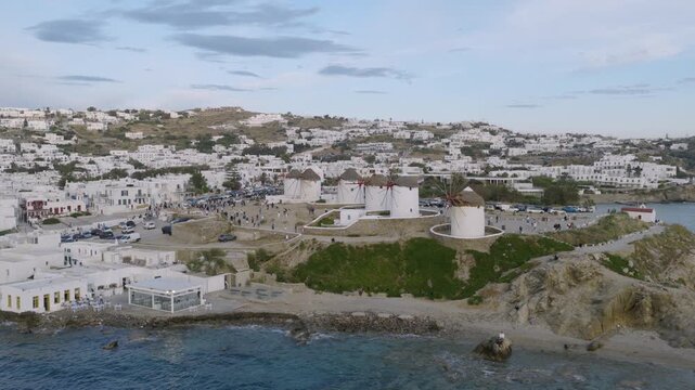 Aerial view of the iconic Mykonos windmills on a green bluff above the beach, with crowds gathered nearby and white buildings covering the hillside.