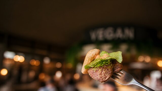 Close-up of a delicious plant-based vegan burger bite held on a silver fork, featuring fresh lettuce and savory patty in a cozy restaurant setting with bokeh background for healthy food concept