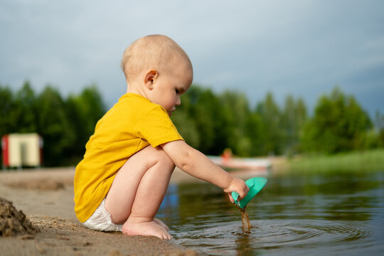 Little boy squats at water edge holding green toy shovel with focused intent. Curious youngster explores sand and water with concentration on summer day. Child discovers nature through outdoor play
