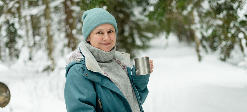 Smiling woman lady holds a silver cup enjoying a hot drink in a snowy forest. Person in winter jacket with fur hood savors a warm beverage outdoors. Adult beams at camera amid snow covered trees