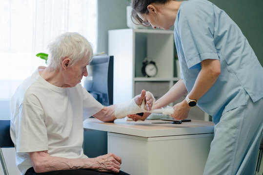 Nurse removing bandage from elderly man injured hand in clinical setting. Healthcare professional carefully unwrapping dressing and providing wound care procedure for senior patient
