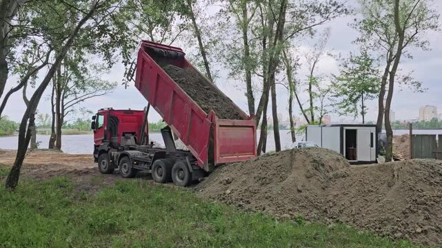 Kyiv, Ukraine may 12, 2026 A horizontal shot showing a heavy red tipper truck unloading pile of soil at an outdoor development site near a riverbank, surrounded by trees and a small construction cabin