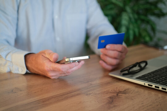 Adult man holding credit card and smartphone while sitting at a wooden desk with laptop and paper.
