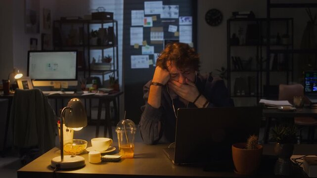 Caucasian young adult man working late on laptop in dark office, yawning and feeling tired. Ideal for commercial themes of corporate burnout, overtime stress, or software development