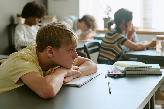 Teenage students studying in classroom, Caucasian boy resting on desk while diverse classmates working behind, showing school focus and fatigue. Useful for education campaigns