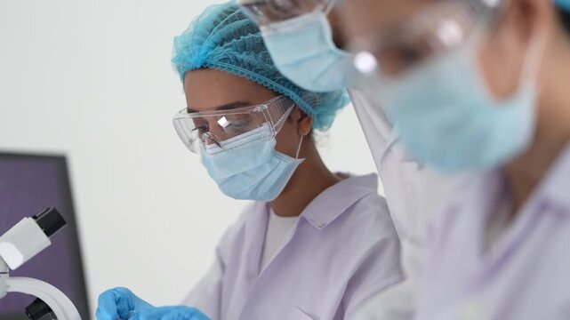 Masked scientists in surgical caps work side by side at a microscope, examining specimens in a bright biology research laboratory.