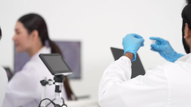 A female scientist with a ponytail and safety glasses smiles while discussing results with a colleague at a computer workstation with a microscope.