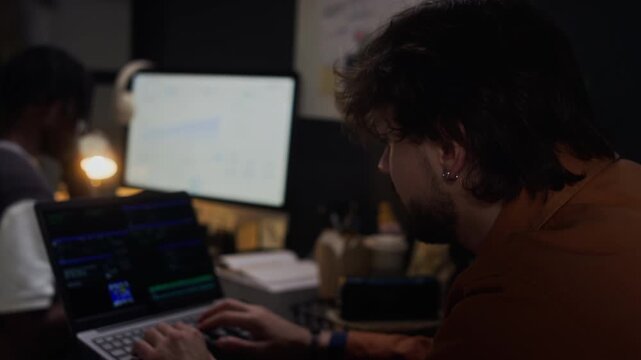 Caucasian young adult male programmer typing code on laptop while African American colleague works in background. Video is suitable for software development and IT startup commercials