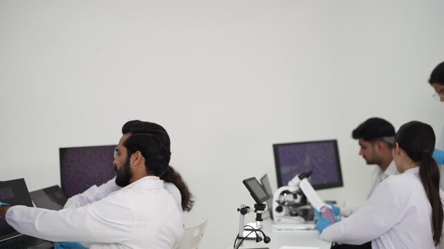 A seated scientist reviews an imaging printout while a female colleague holds a tablet beside a microscope and DNA graphics on screen.
