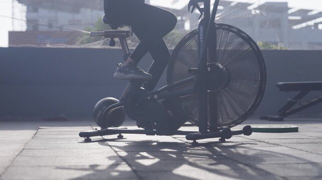 Tracking shot of the lower body of a South Asian female athlete pedaling an assault air bike during functional fitness workout in a modern indoor fitness studio. Collection: WORKOUTSERIES2026.
