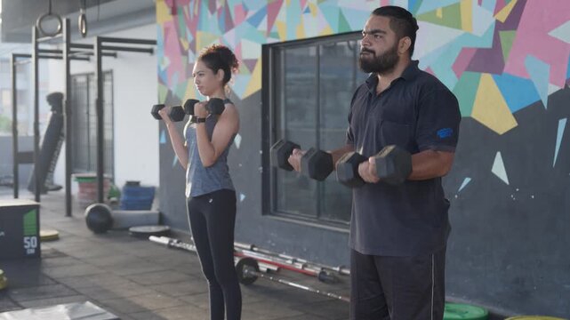 South Asian male and female athletes perform standing dumbbell bicep curls in a modern indoor gym. Tracking shot with push in and pull out camera movement. Collection: WORKOUTSERIES2026.