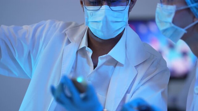 A masked scientist examines small blue sample capsules under dramatic blue laboratory lighting with molecular graphics on the monitor.