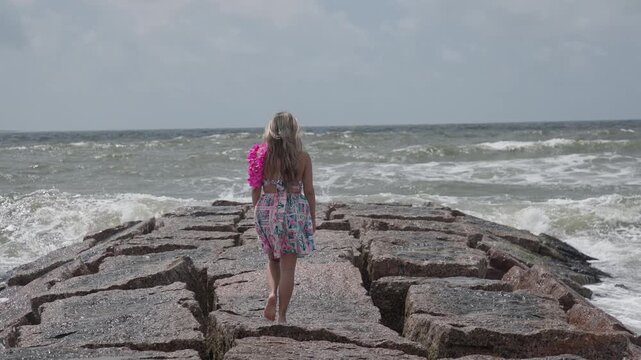 Blonde girl in summer dress and pink float walks barefoot on a rugged rock jetty at Galveston Beach as dramatic waves crash all around her.