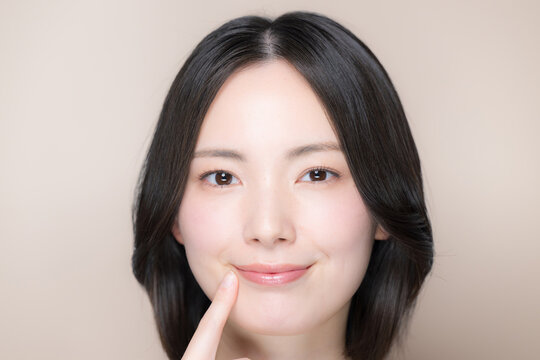 A Japanese woman facing the camera against a pink-beige background, smiling and showing her beautiful mouth and lips&mdash;a close-up beauty portrait with natural makeup, pointing to her mouth with her fing