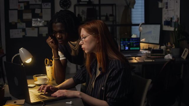 Young adult Caucasian woman and African American man collaborating on laptop in office at night. Ideal for corporate training, teamwork concepts, or software development marketing