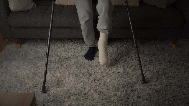 Person with a broken leg in a cast using crutches to carefully stand up from a sofa at home. Injury recovery, rehabilitation, limited mobility and physical therapy concept.