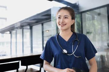 A smiling Asian female doctor or nurse in a blue uniform with a stethoscope around her neck is...