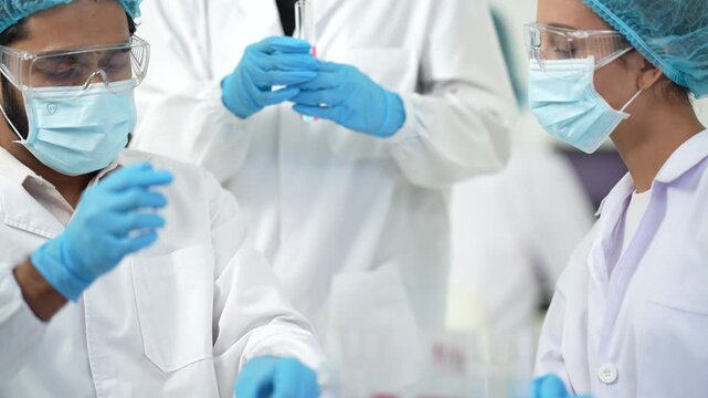Two masked scientists handle a small sample tube with gloved hands, one giving a thumbs-up in a bright research laboratory.