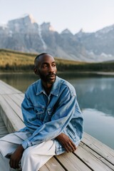 Fototapeta premium Thoughtful man seated on lakeside dock wearing denim jacket by mountain water for lifestyle background and banner design