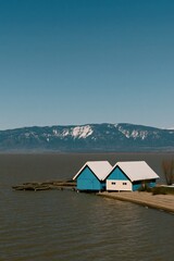 Fototapeta premium Blue boathouse duo on a calm lake with snow-capped mountain background for lakeside wallpaper and banner design