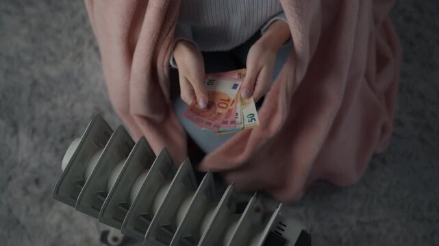 Woman struggling to stay warm at home while holding her hands over a radiator with euro banknotes in hand . Cost of living crisis, inflation, rising household expenses and financial stress concept.